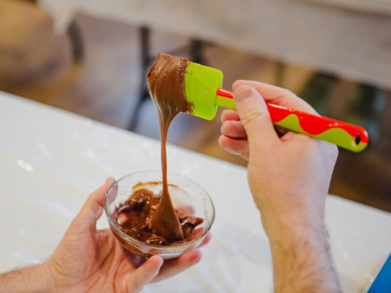 A green spatula with chocolate pouring from it into a bowl